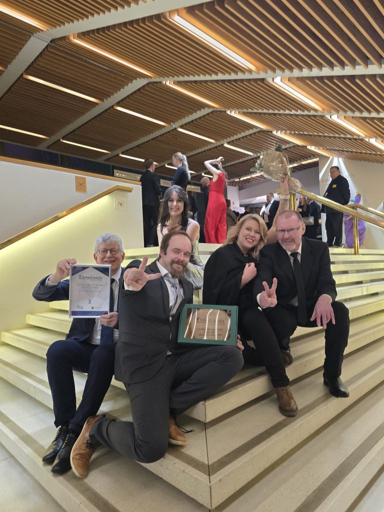 Kuvassa portailla istuvat UKKIEN pelaajat Kalle Kärkkäinen, Ville Korhonen, Anfisa Kolesnik, Mona Koskelo ja Keijo Korkalainen. Pelaajat hymyilevät ja tuulettavat.