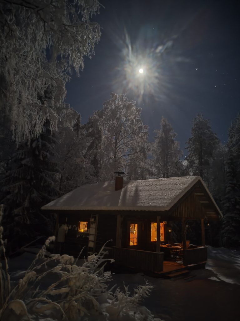 Kuvassa on öinen maisema, jossa erottuu kuunvalo ja pieni mökki, jonka ikkunasta kantautuu ulos lämmintä valoa.