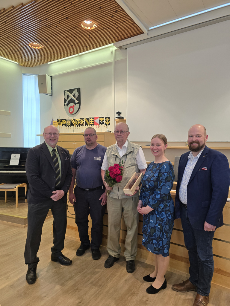 Kuva valtuustosalista, jossa seisoo vierekkäin viisi ihmistä. Kunnanvaltuuston puheenjohtaja Ilkka Roininen, kunnanhallituksen puheenjohtaja Timo Tuovinen, pitäjänneuvos, valtuutettu Eero Kukkonen, hallintojohtaja Maria Puustinen, kunnanjohtaja Mikko Kärnä.