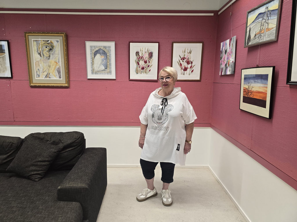 Kuvassa Merja Hanhinen poseeraa taidenäyttelyn avajaisissa. Taustalla useita Merjan maalauksia, jotka on ripustettu kirjaston seinälle.